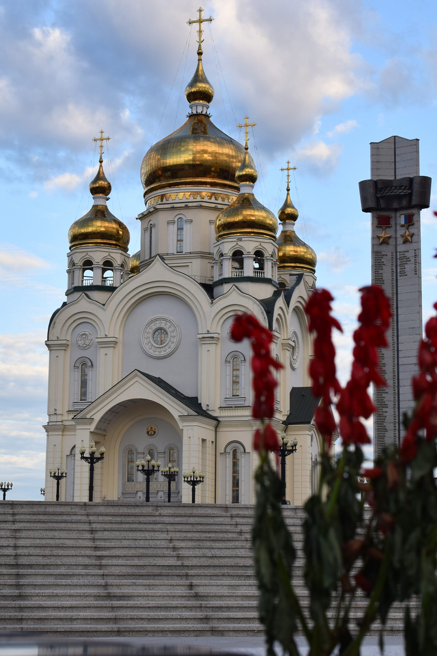 White massive cathedral with five gilded domes on the square, obelisk and stairs in front of the cathedral, red flowers in the foreground
