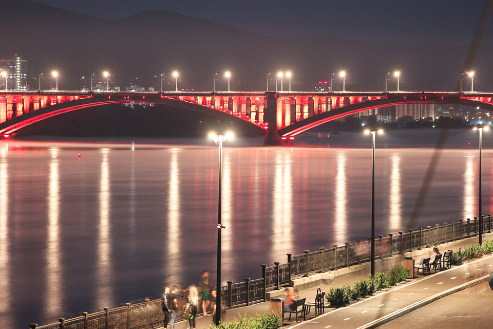 A bridge illuminated at night over a wide river, river embankment with people walking, symbol of the city