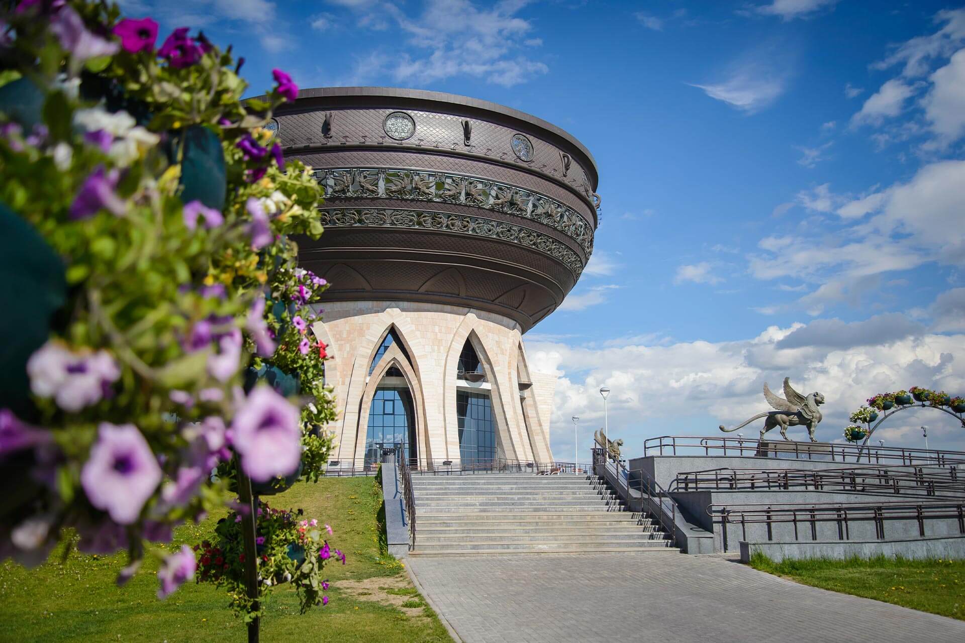 Round building of an unusual architectural style. The base of the building with gothic arch shaped windows. The top is in shape of a bowl or a copper. Flowers in front of the building