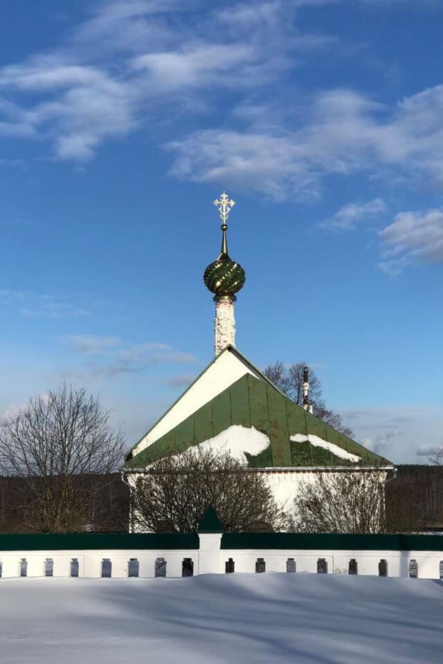 A white brick monastery wall with a green roof and a dome crowned with a golden cross in winter.