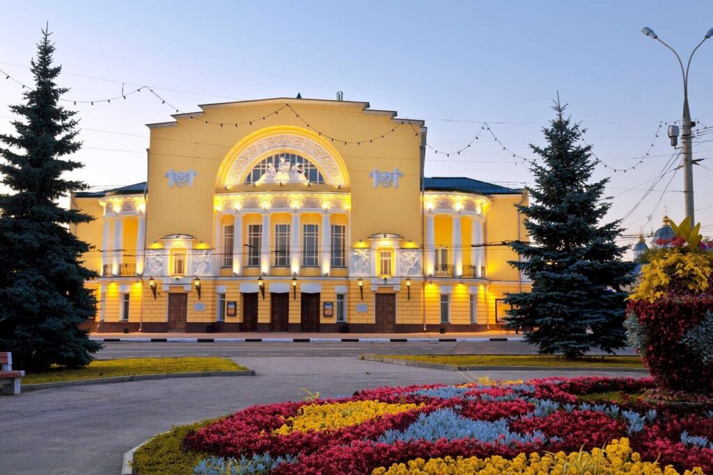 A yellow classical building with white columns, façade decorated with a sculpture. Colorful flowerbed in front of the theatre .