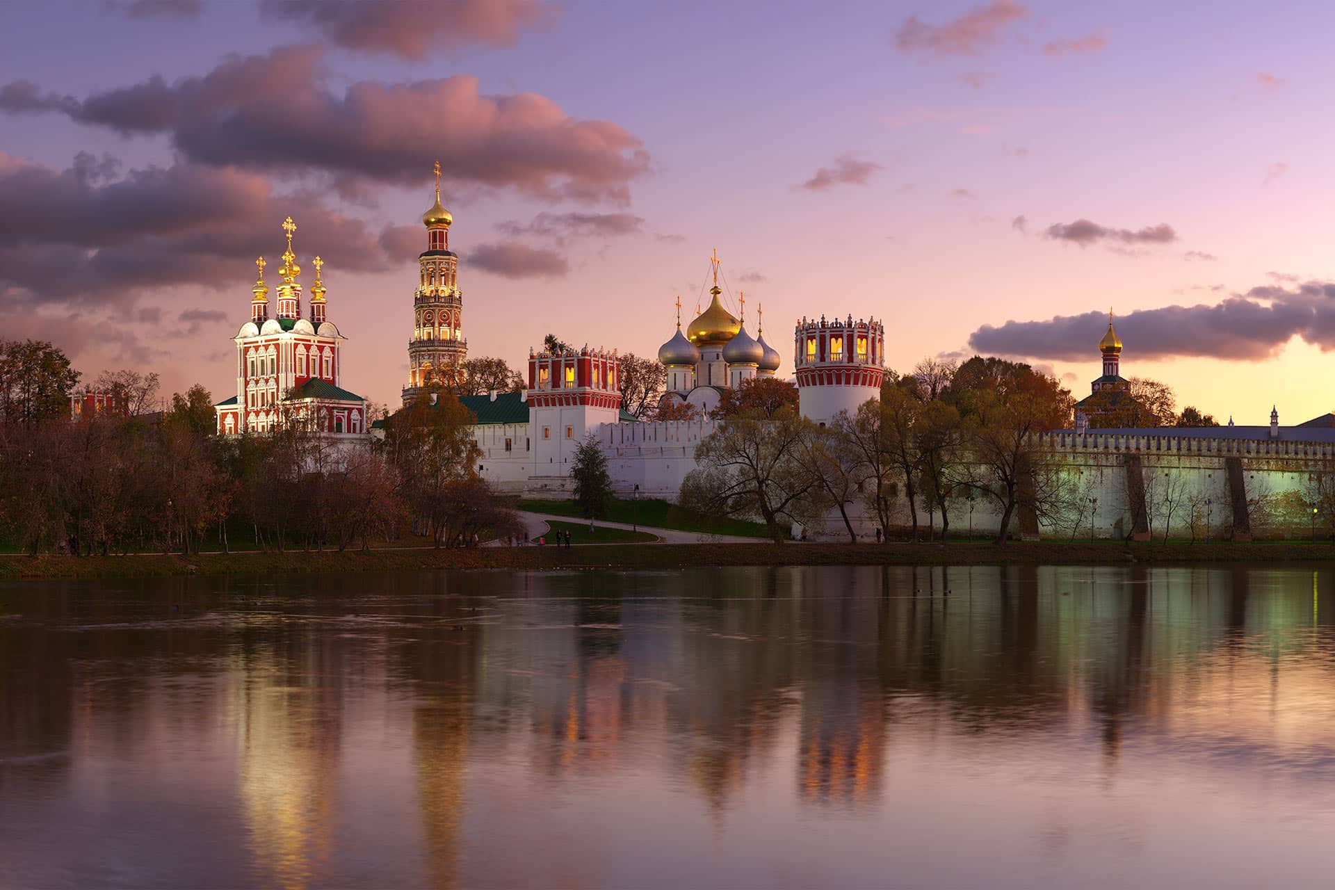 An architectural ensemble of several orthodox churches behind the white wall on a river bank in the evening