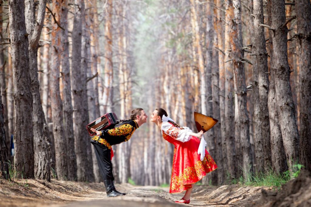 A man and a woman wearing traditional Russian clothes with traditional Russian musical instruments accordion and balalaika in birch forest going to kiss