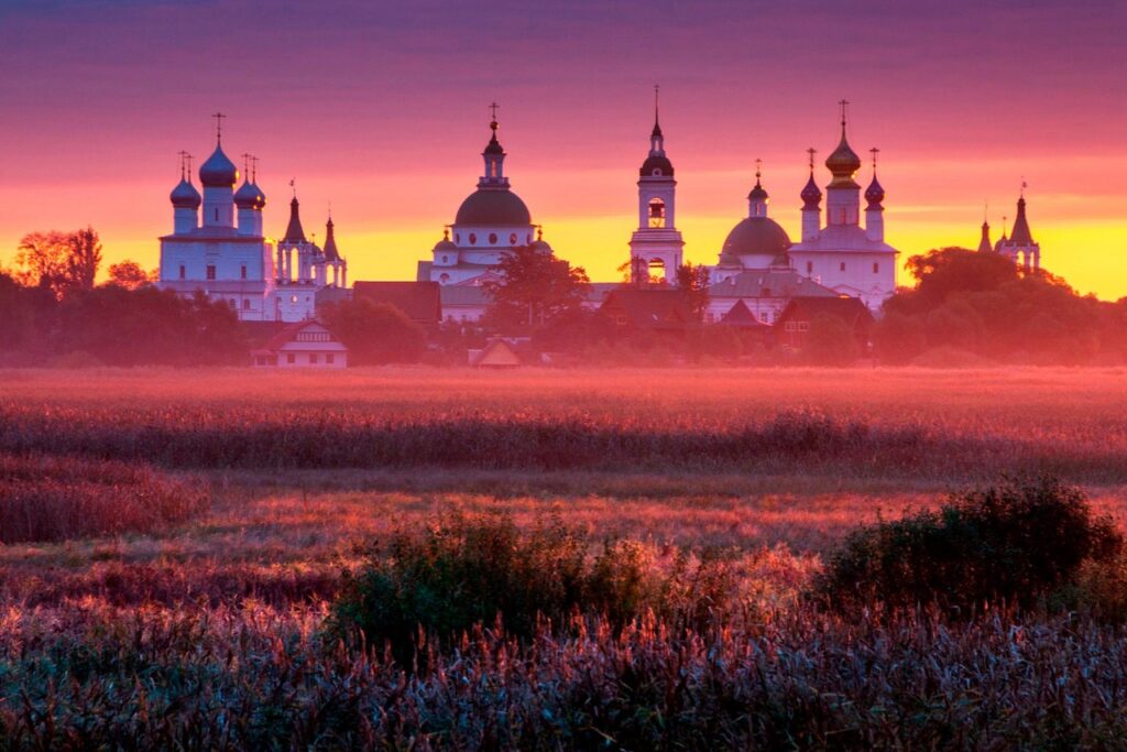 A field in front of a monastery with many churches with domes of different colors during sunset