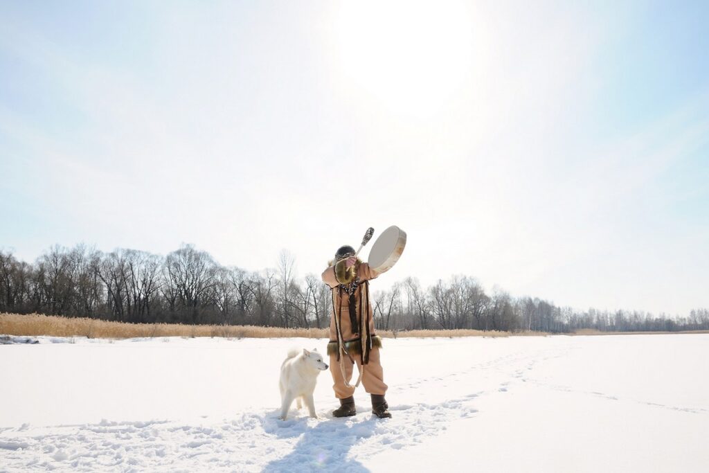 A Shaman with a tambour and his dog in a winter landscape