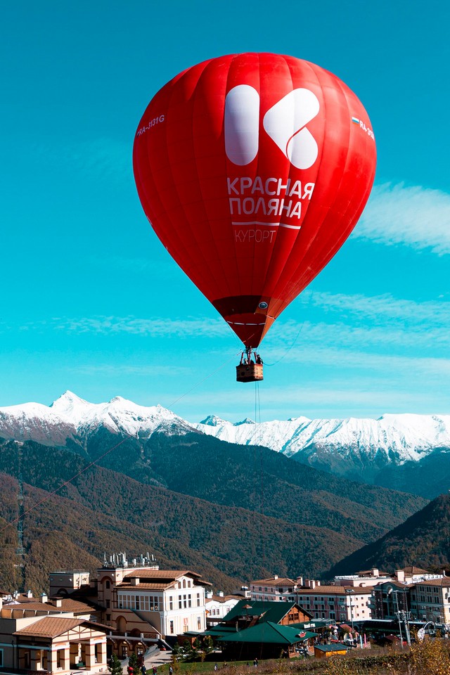 A red hot air balloon with a sign “Krasnaya polyana” in Russian, flying over the mountains and village