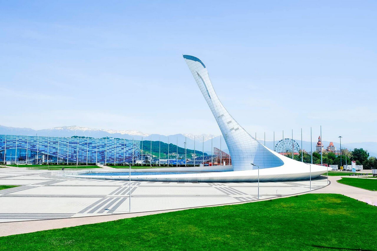 Olympic fire bowl in the shape of a wing holding a pool with fountains, blue stadium and mountains in the background