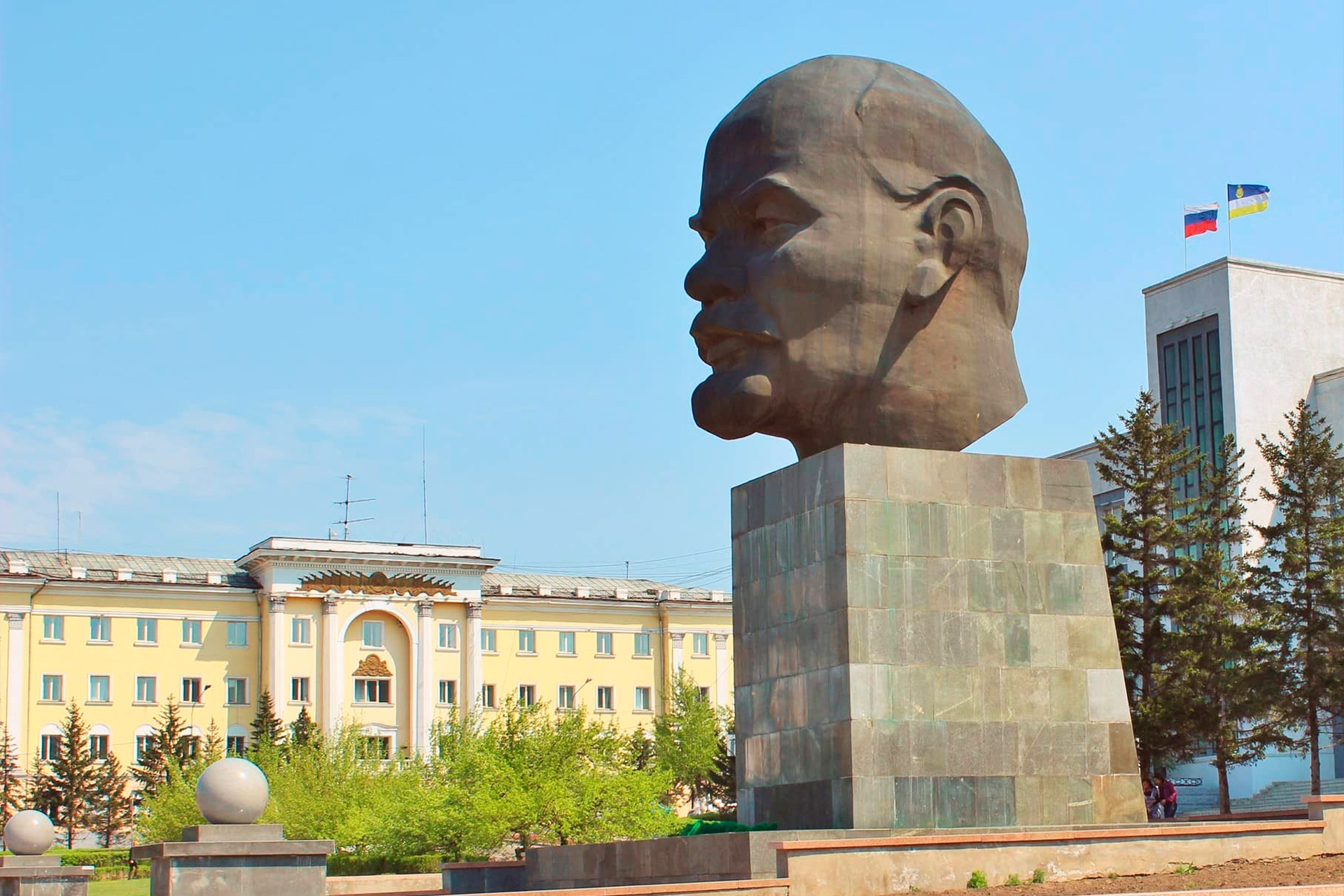 A grey pedestal with a giant head of a man on it. White and yellow buildings are in the background.