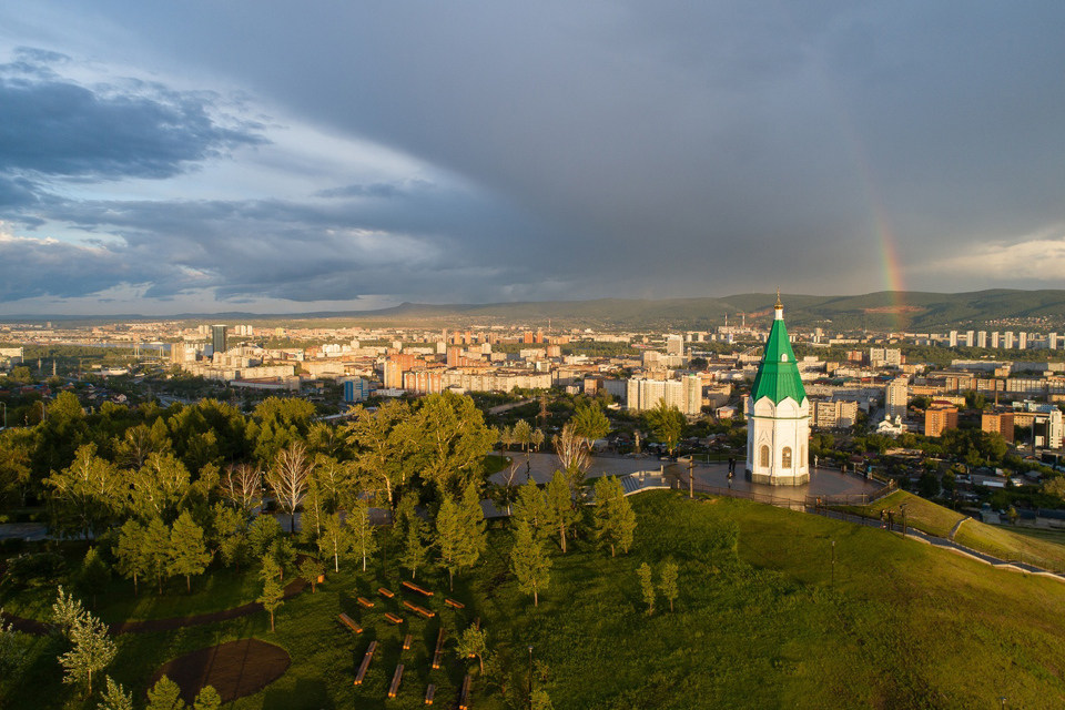 A small white orthodox chapel with green peaked roof on a hill, a panorama of a big city from the hill, rainbow in the sky