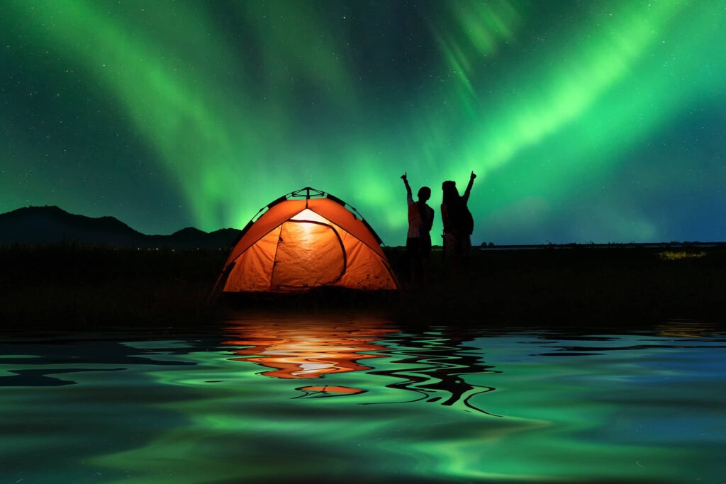 Two people near a tent watching the green northern lights in the sky