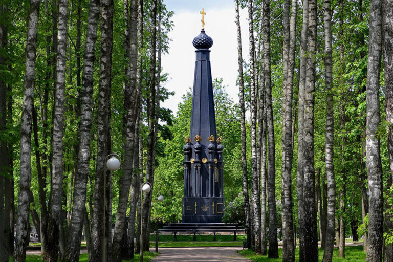 A memorial in a park looking like a black chapel with an orthodox cross at the top and small decorations in the shape of onion-domes topped with gilded two-headed eagles around the main part of the monument