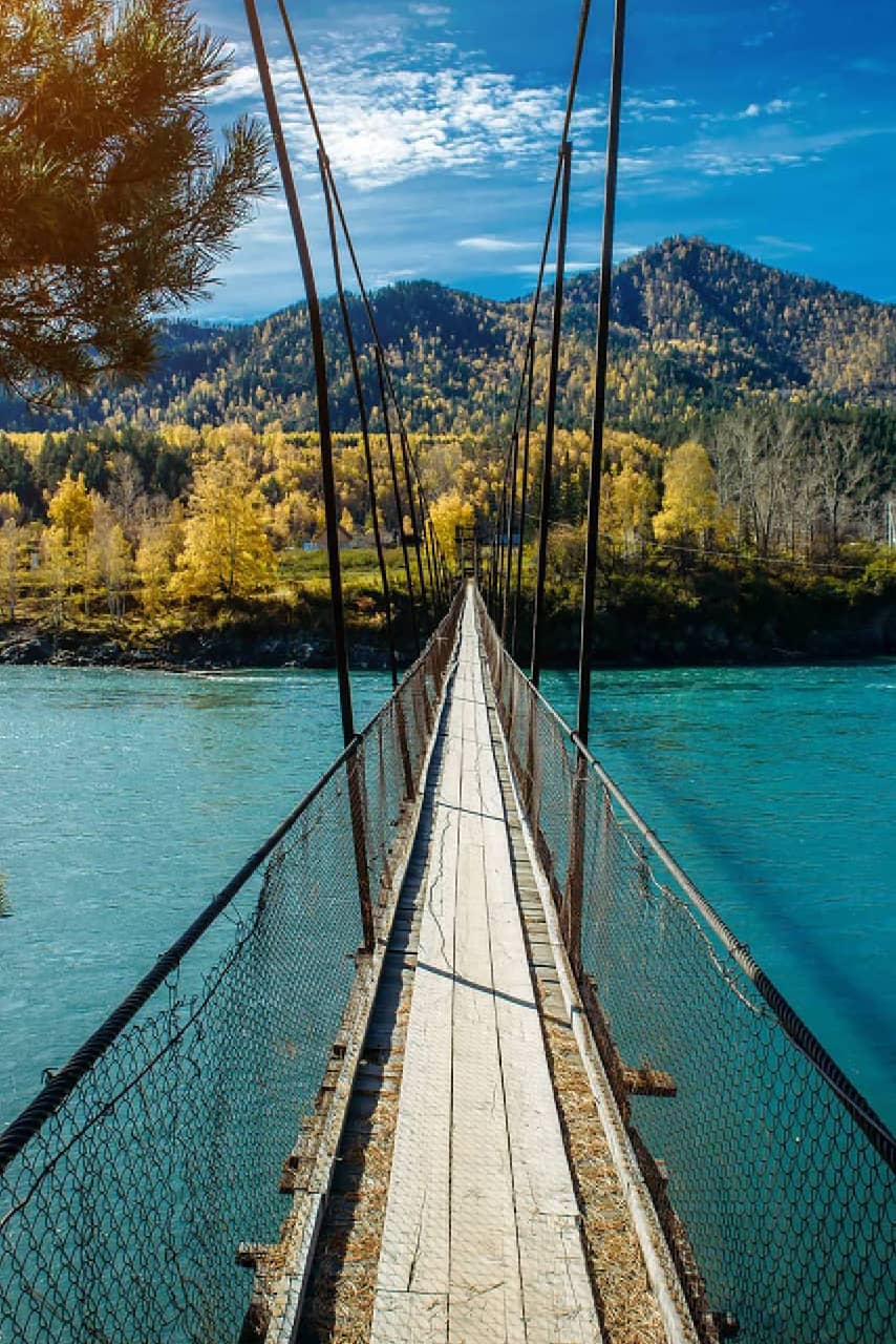 An old wooden pendant bridge over a blue river, mountains and yellow trees on the other side of the river