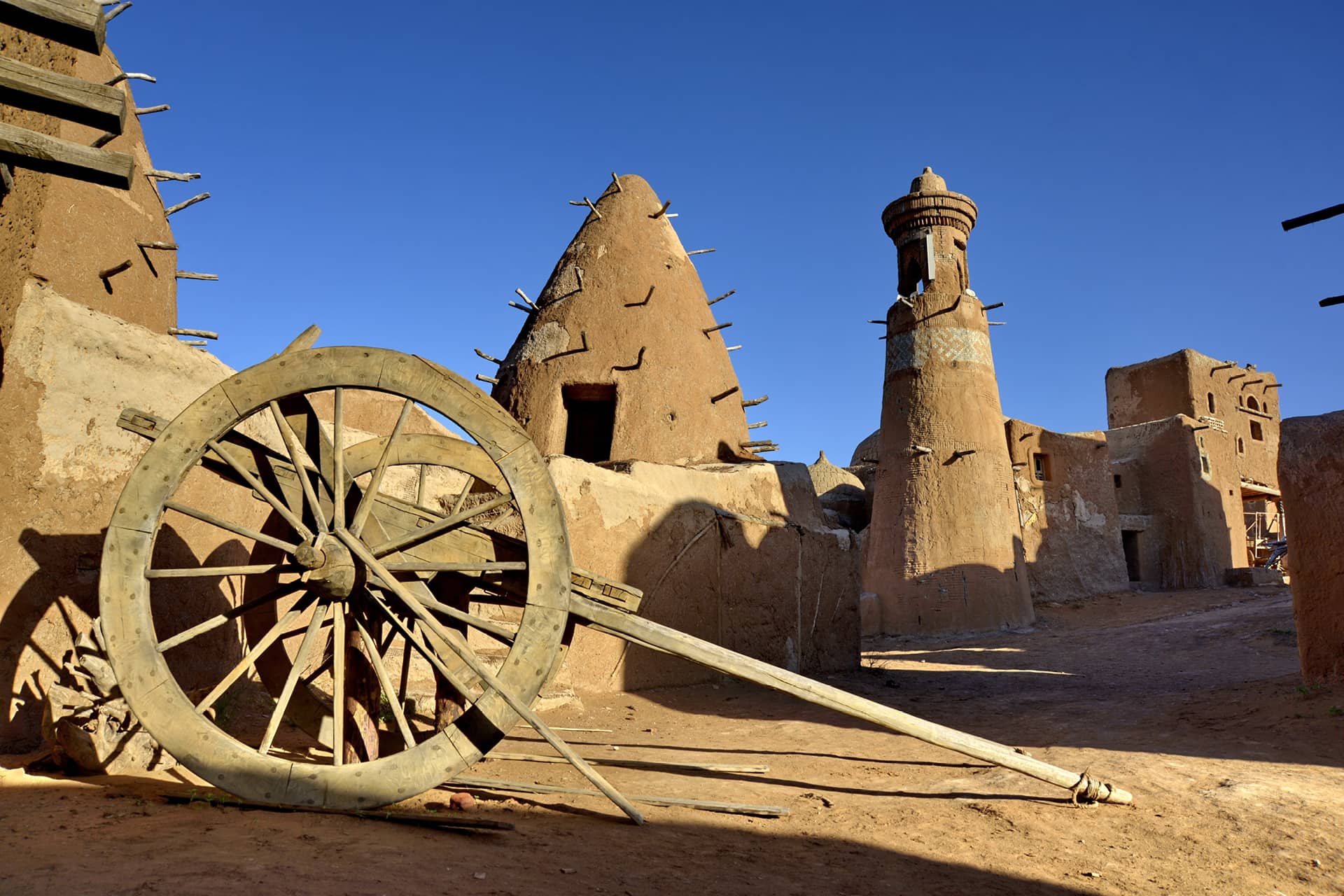 Buildings of an old ruined city-museum in the Middle East, a brown fortress wall with cone-shaped towers with wooden support sticks inside, a lighthouse, a wooden wagon in front.