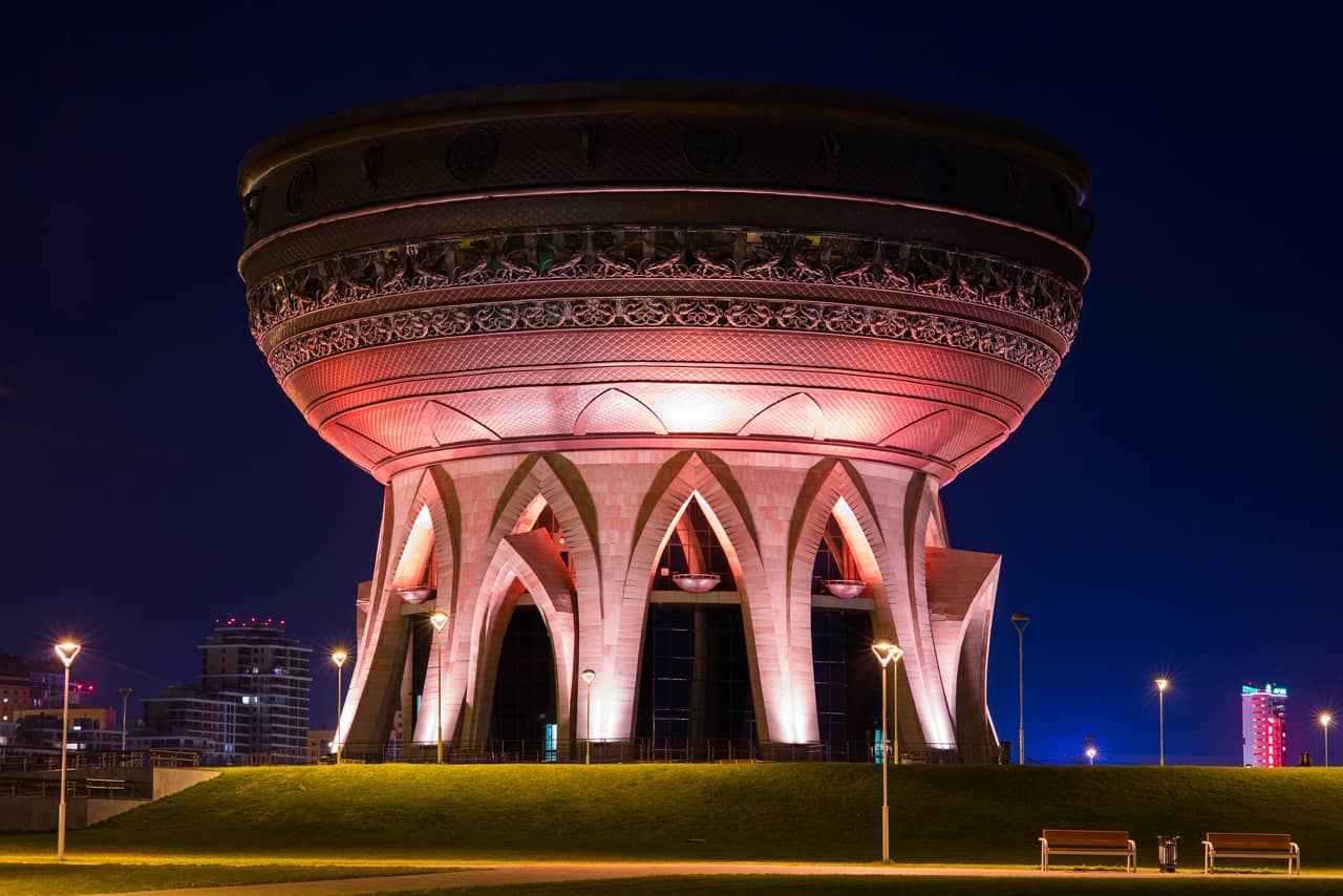 Round building of an unusual architectural style at night. The base of the building with gothic arch shaped windows. On the top there is a double-story observation deck on the roof in shape of a bowl or a copper .