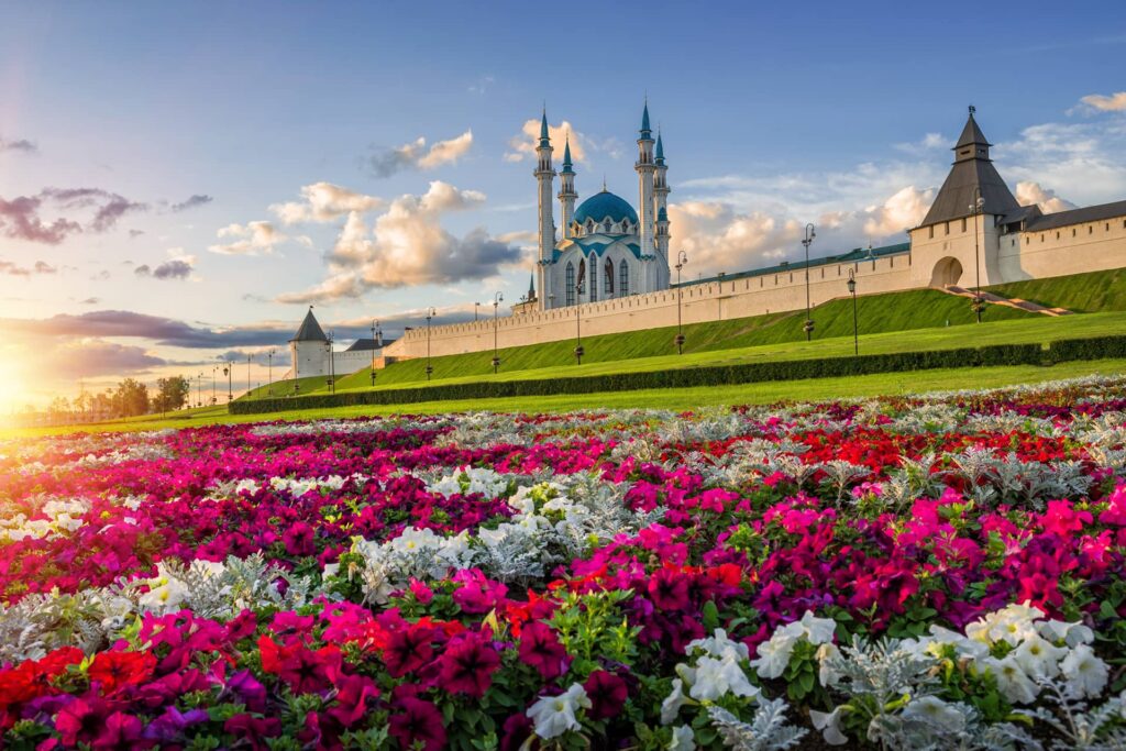 A colorful flowerbed in front of a fortress and a white mosque with blue domes and four minarets in the distance