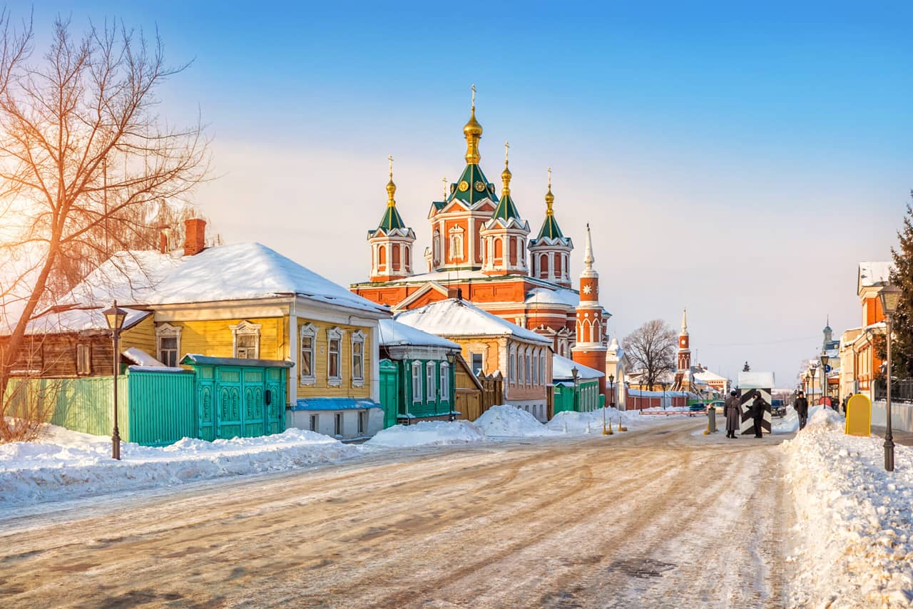 A snowy street of small Russian town, colorful wooden houses and a small red church in Kolomna, small red church with hexagonal towers and gilded domes