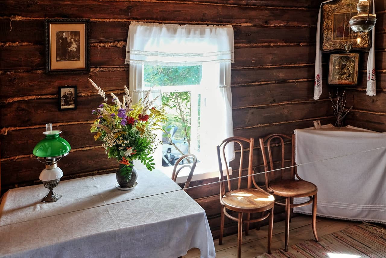 A room in an old wooden house that is now a museum, a window with white curtain, table covered with white tablecloth and flowers on it, portraits on the walls and an icon in the corner of the room