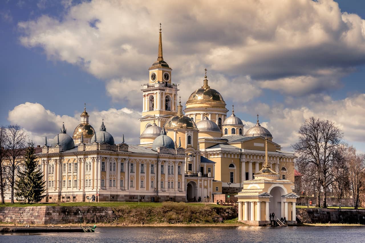 A complex of five yellow and white churches of neoclassical style with gilded domes, a bell tower with a clock, monastery on a shore of a lake, portico with a steeple on the top on the lake in front of the monastery