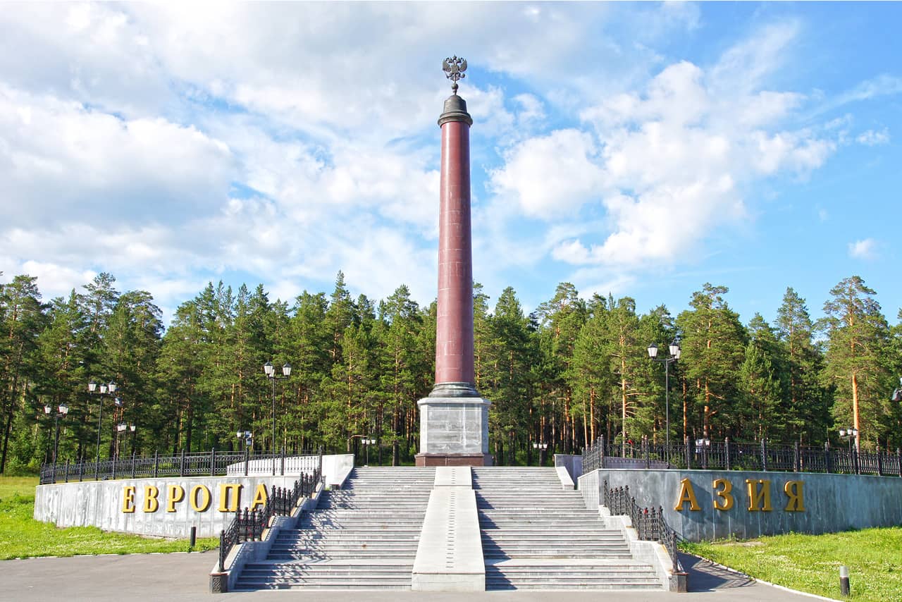 A marble obelisk with a two-headed eagle on the top on a square in a forest, stairs in front of the obelisk, signs in Cyrillic saying “Europe” and “Asia”