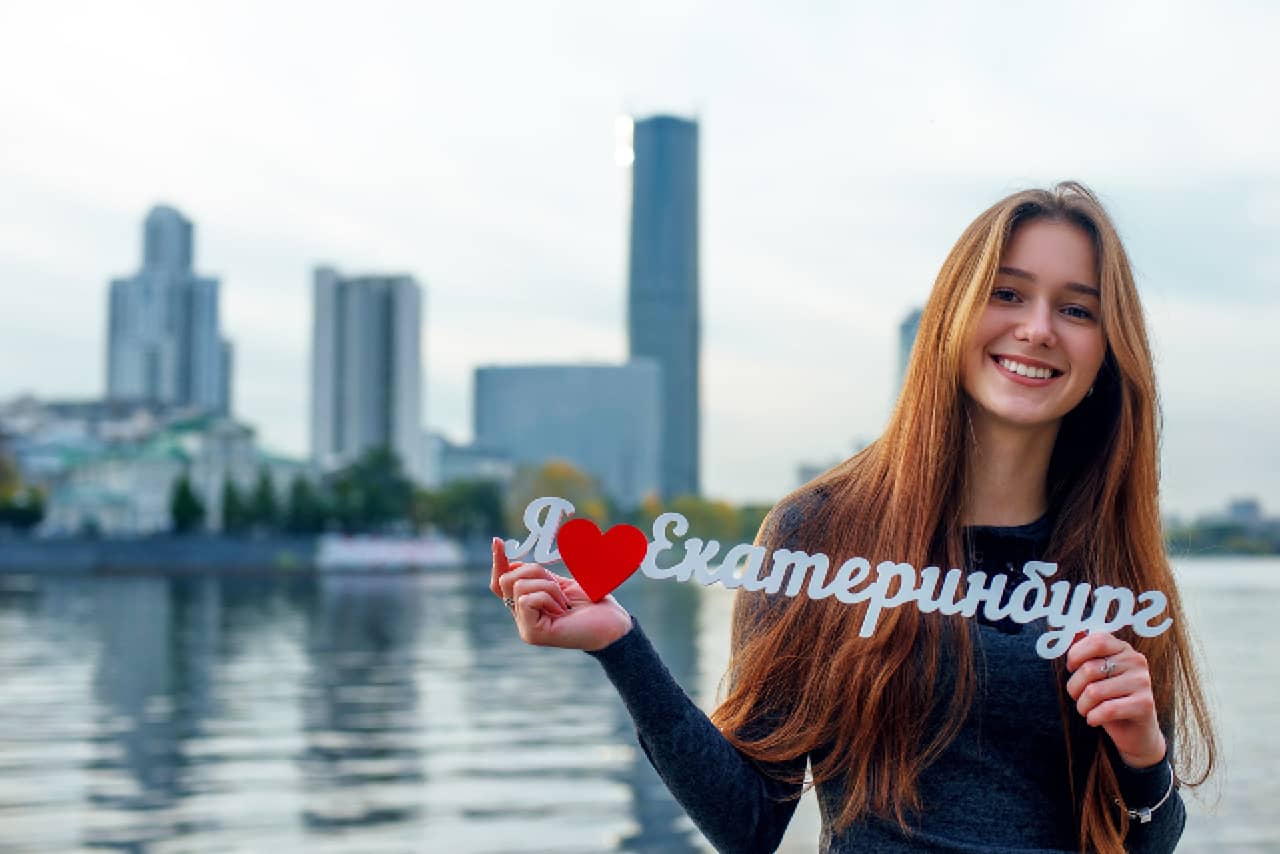 A young lady with long hair in front of a river and modern buildings, a sing in Russian language “I love Yekaterinburg” in her hands
