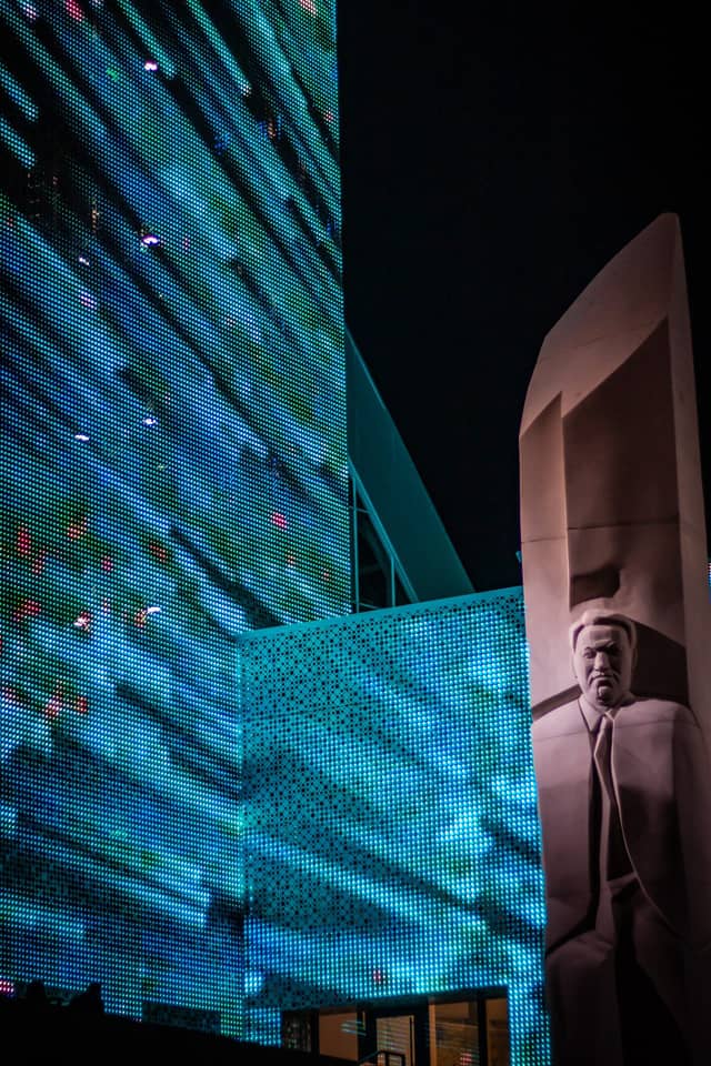 A modern building illuminated at night, all the wall of the building is a large screen with blue lights, a fresco of a man on a wall on the right