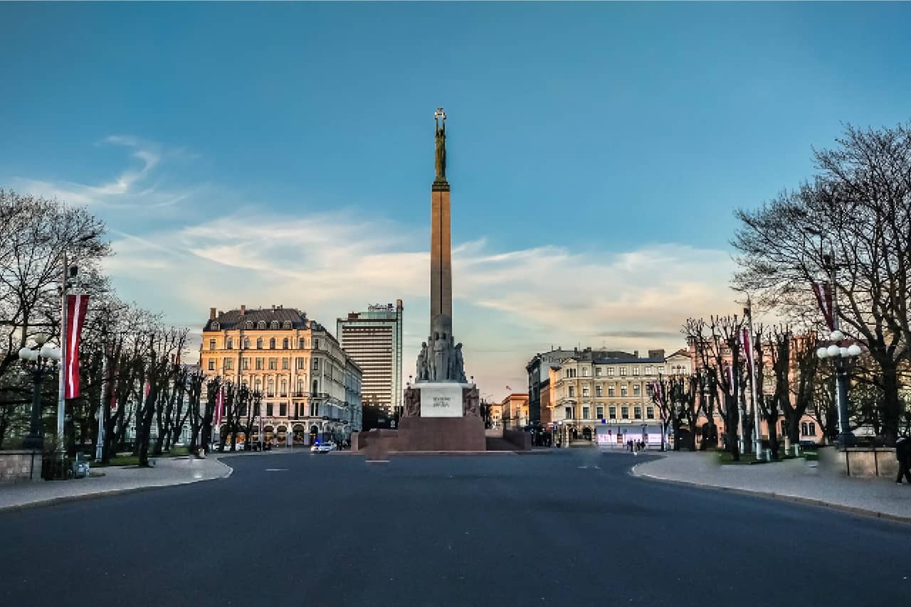 An obelisk with a statue of a woman lifting three gilded stars, monument surrounded with beautiful buildings in classical style and a park