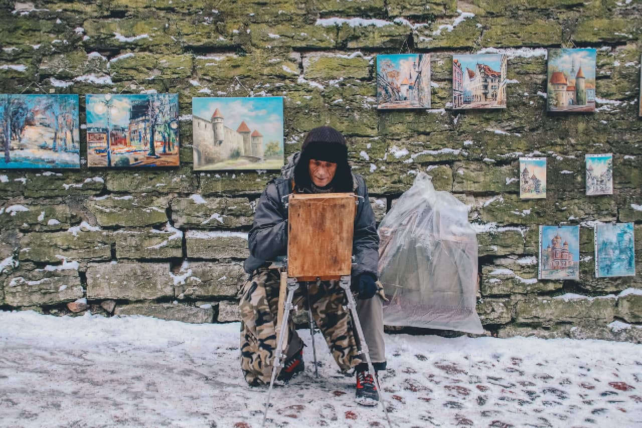 An old man wearing a winter fur hat painting and selling his pictures in the street in winter