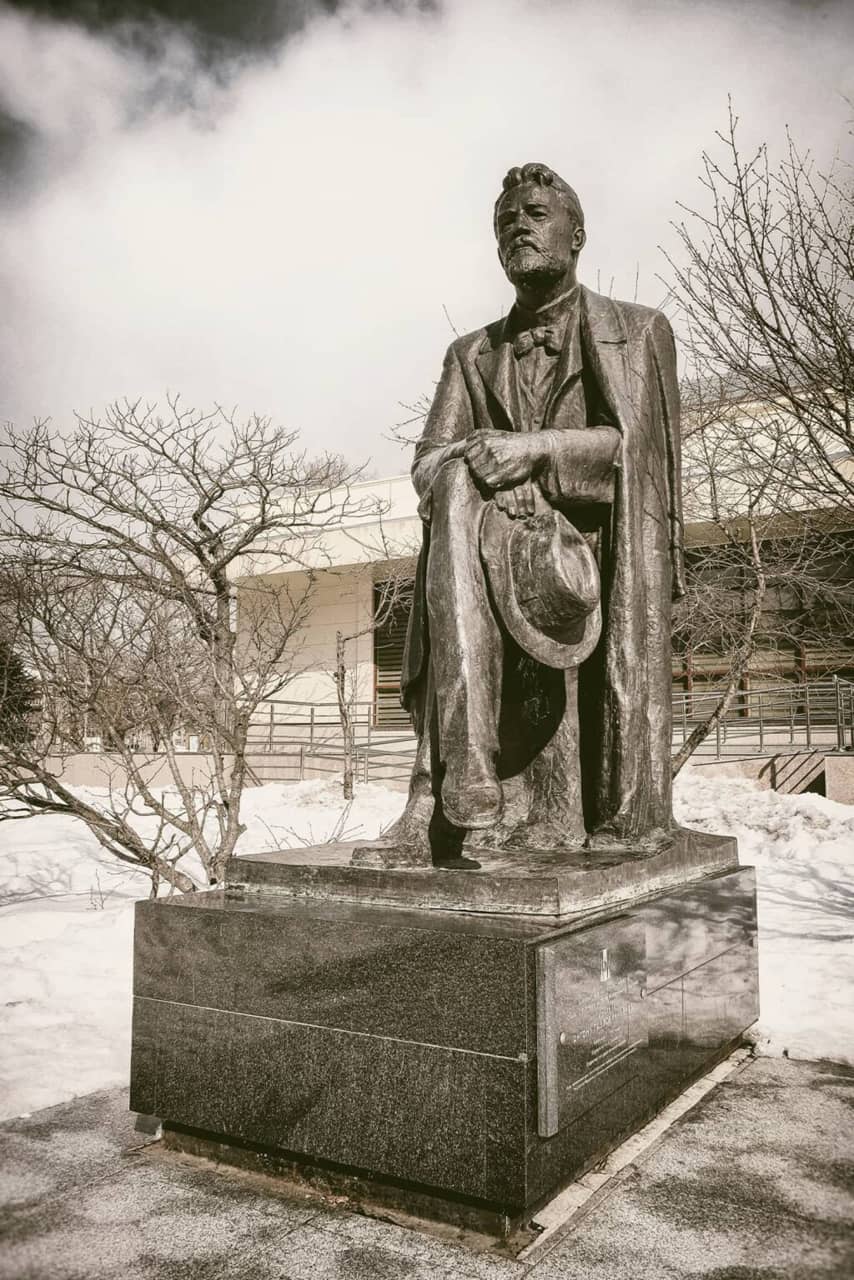 A bronze monument of a sitting man holding a hat in his hands in winter