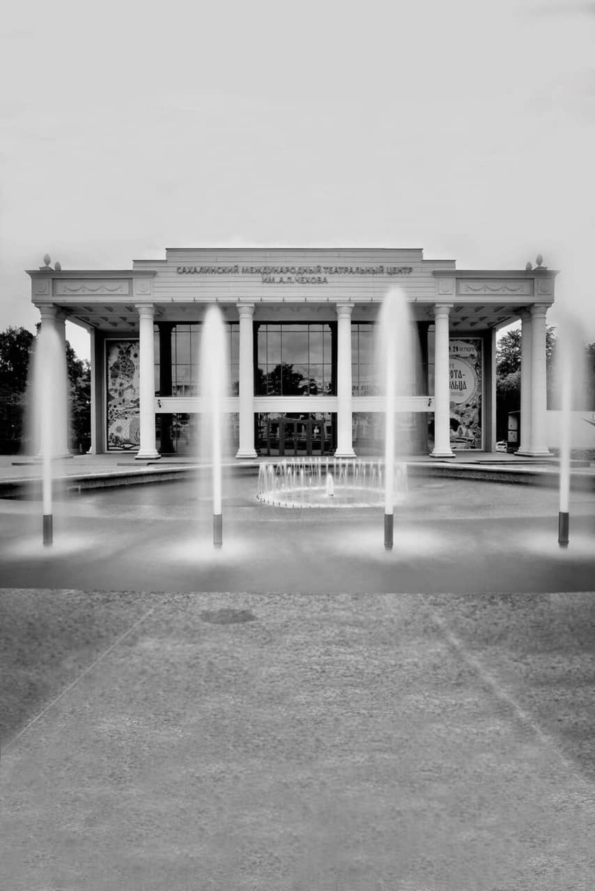A black and white photo of a building with giant windows and portico decorated with columns in Soviet style and waterfalls in front of the building