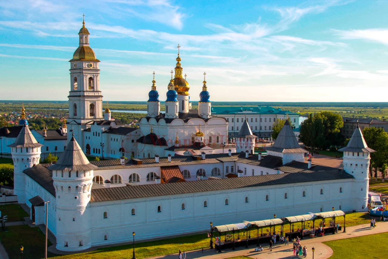 A beautiful renovated complex of a fortress of rectangular shape with round towers and a white cathedral with blue and golden domes behind the fortress, a bell tower