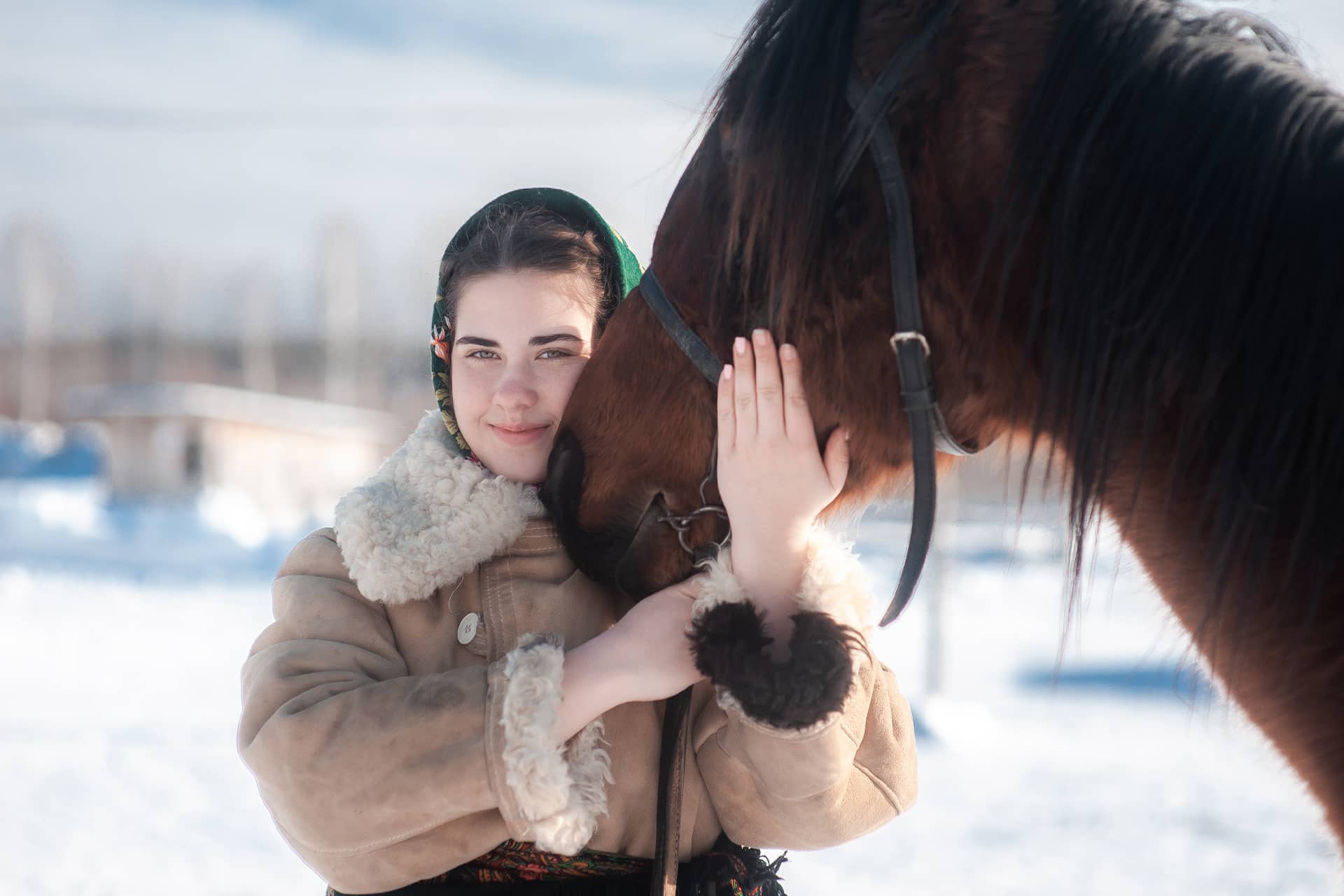 A Russian girl wearing sheepskin coat hugging the head of a horse in winter