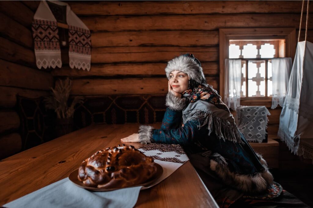 The wooden interior of a house, woman in traditional clothes sitting at the table with a loaf of bread in front of her