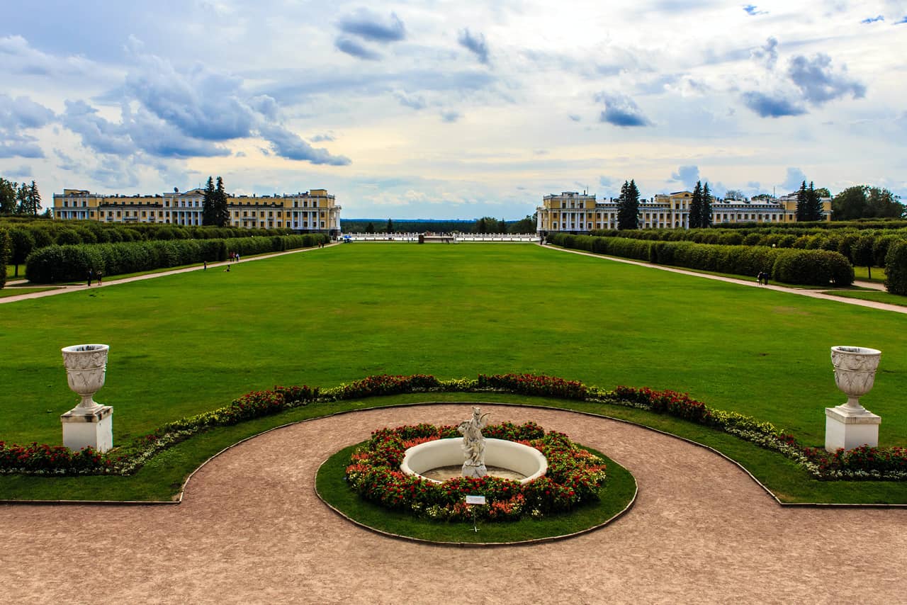 A garden of a palace with a round flowerbed with a white classical sculpture in the middle, two wings of a yellow palace in the distance