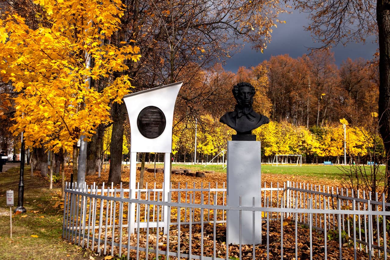 A portrait sculpture of a Russian poet and a memorial next to it, the sculpture group surrounded with a fence, park in autumn, Pushkin’s autumn