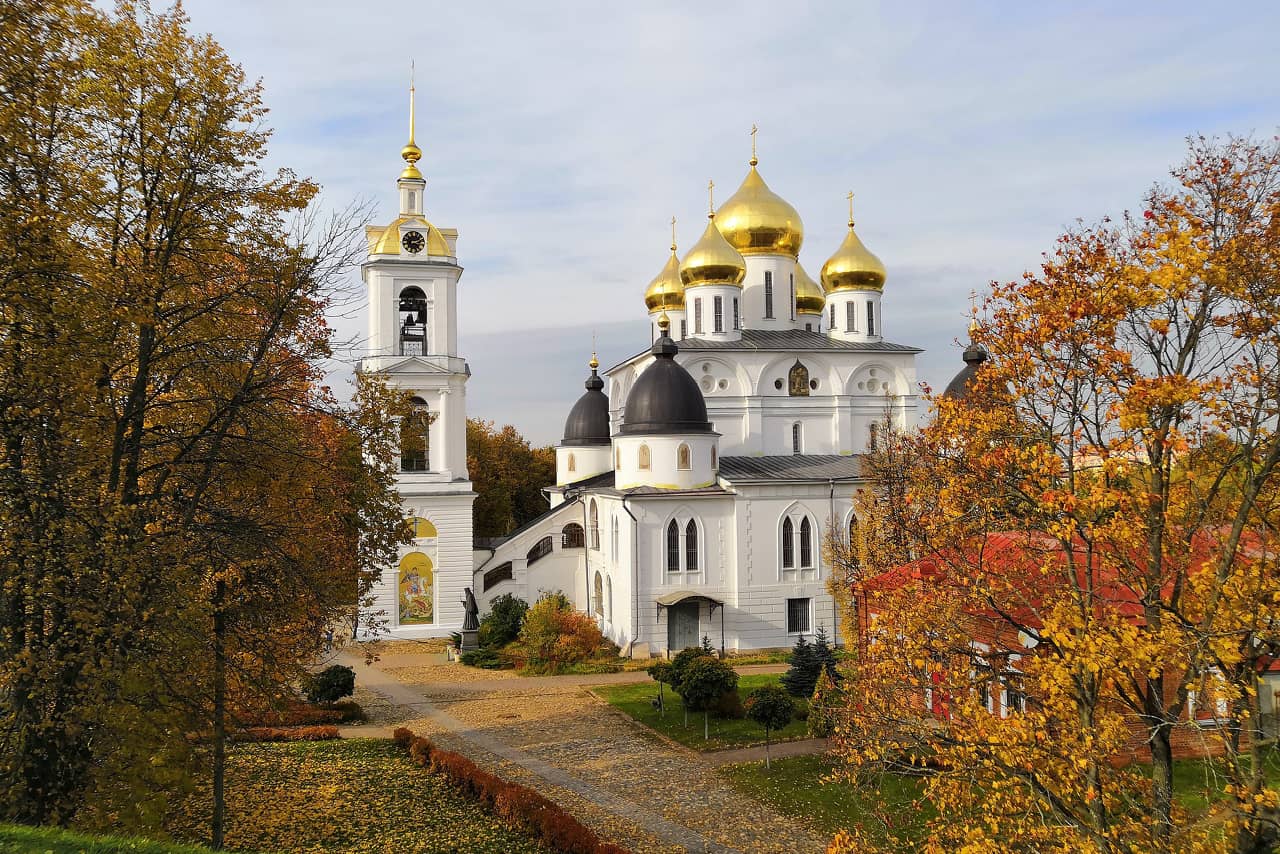 A white church of two levels and a bell tower, an Orthodox church with two round towers topped with black domes and a central part with five golden domes, yellow trees in front of a church